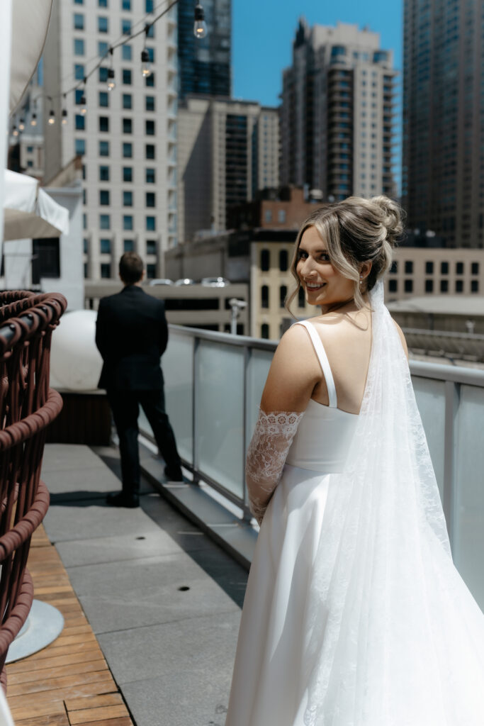 Anticipation builds in this candid photograph of a smiling bride looking over her shoulder toward her waiting groom on an urban balcony just moments before their first look wedding photography reveal.