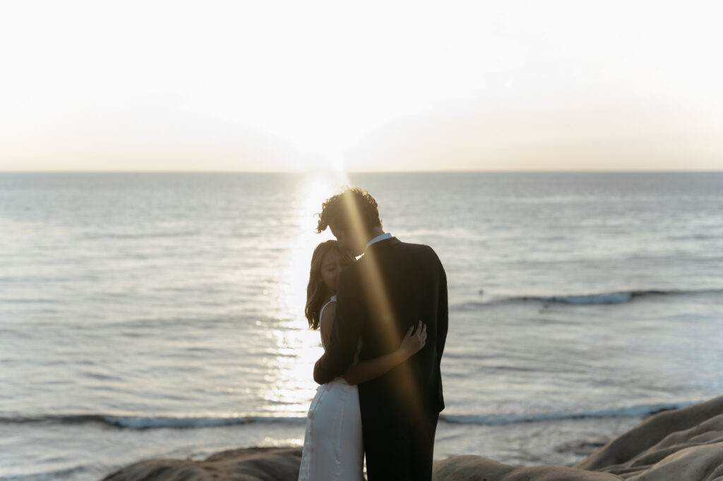 Warm golden sun flare surrounds a couple embracing on the rocks by the ocean during their adventurous location portrait session.