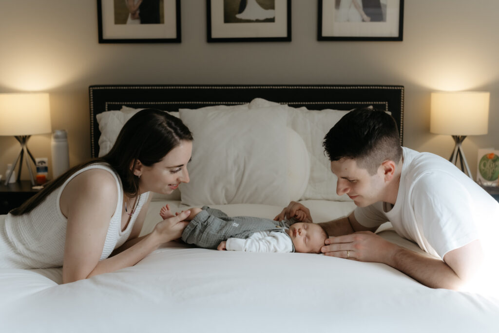 A peaceful lifestyle newborn session featuring parents leaning over their baby on a neutral, sunlit master bed.