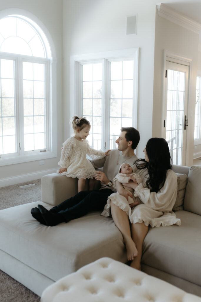 In-home family newborn photography with parents, a toddler in lace, and a newborn baby sitting on a beige couch.