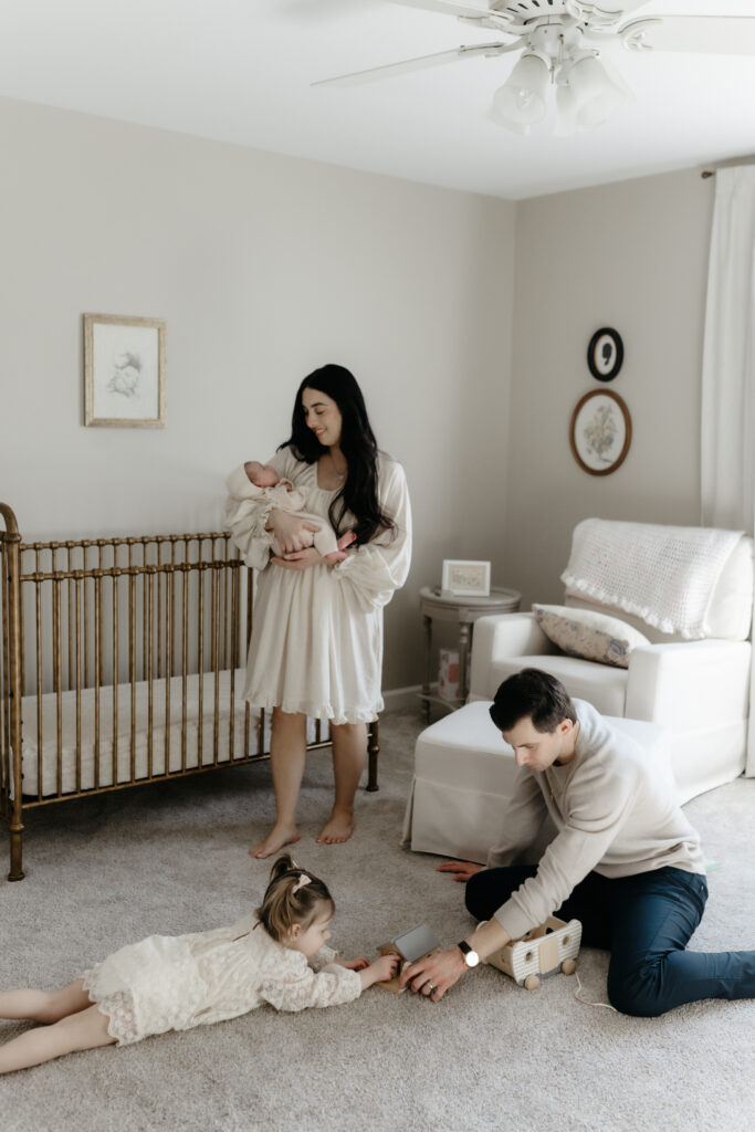 A toddler and father playing with toys on a nursery floor during an in-home family newborn photography session while the mother holds the baby.