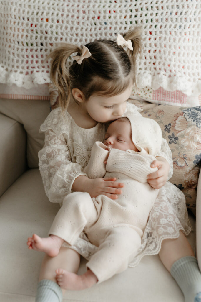 An older sister with pigtails kissing her sleeping newborn baby sibling on the forehead during a family newborn photography session.