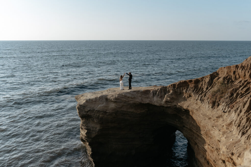A wide landscape view of a couple holding hands and dancing on a dramatic sea arch cliff above the ocean during an adventurous location portrait session.
