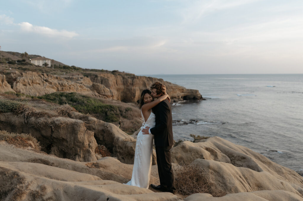 The groom kisses the bride's forehead as they stand on rugged coastal cliffs during an adventurous location portrait session at sunset.