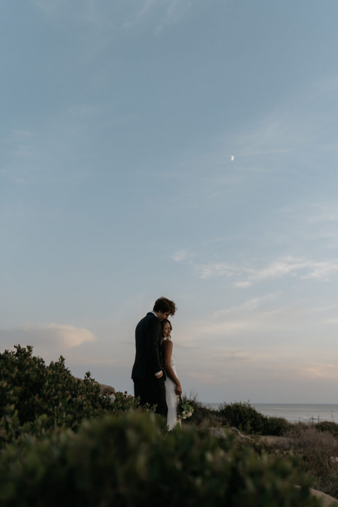 A bride and groom share a quiet moment on a grassy cliffside beneath a crescent moon during an adventurous location portrait session on the coast.