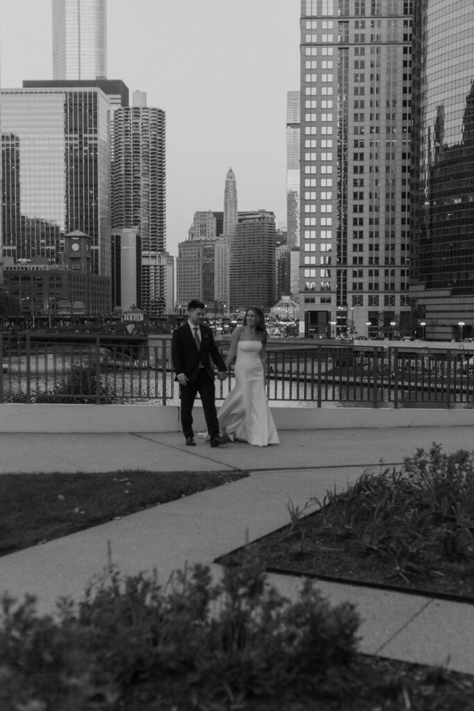 A couple walks hand-in-hand along a city bridge with the towering architecture behind them.