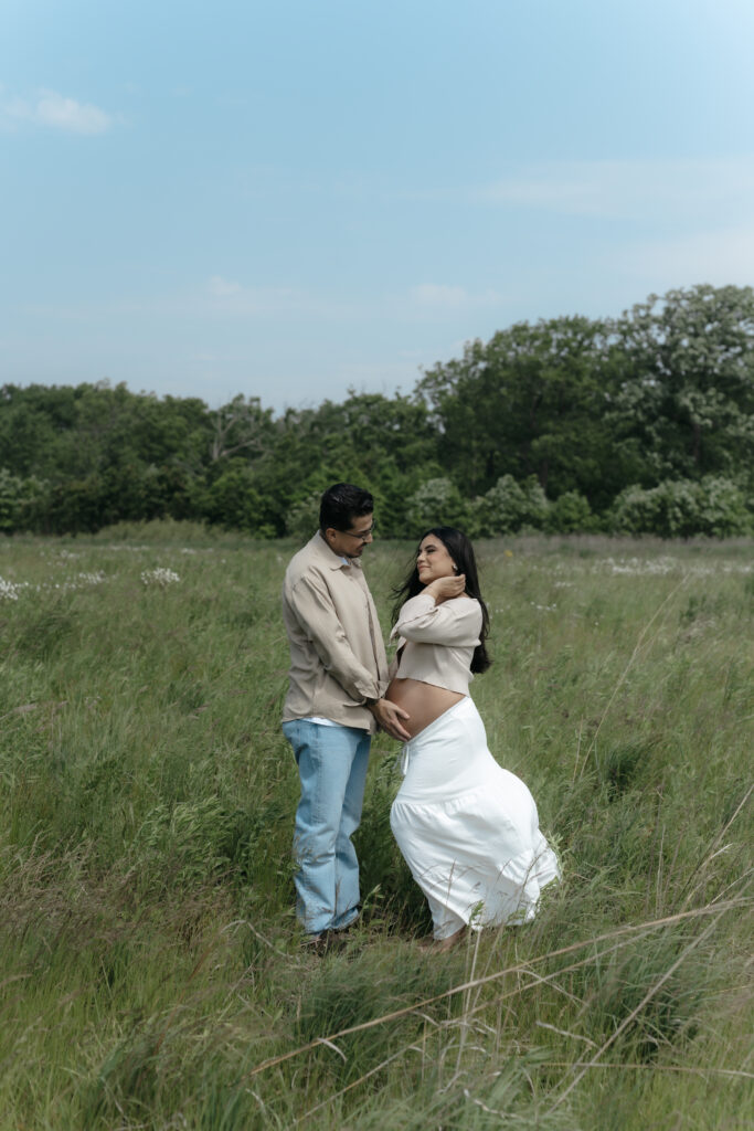 A couple posing together in a windy field wearing neutral linen and denim maternity clothes for photoshoot day.