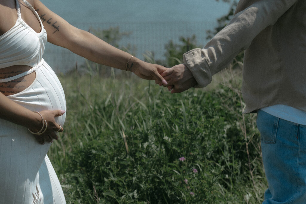 Close up of a couple holding hands outdoors showing a white maternity dress and a tan linen shirt.