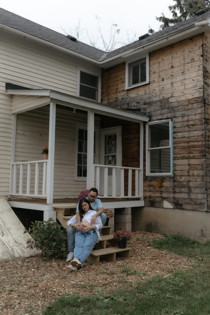 A happy couple cradling a pregnancy bump while sitting together on porch stairs surrounded by autumn flowers.