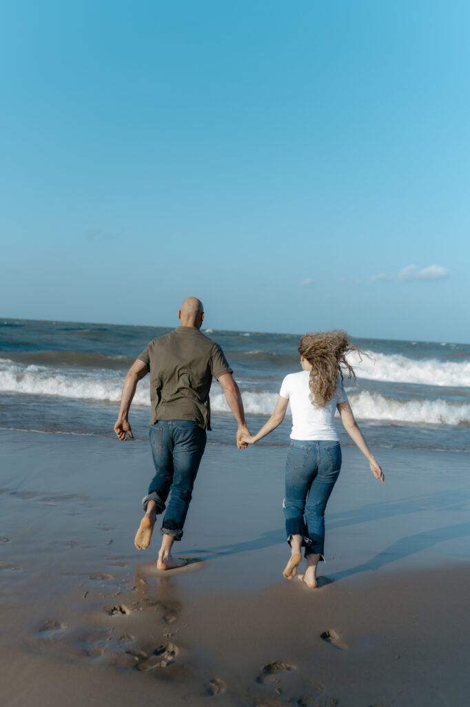 A couple running barefoot toward the ocean waves exploring fun engagement photo ideas.