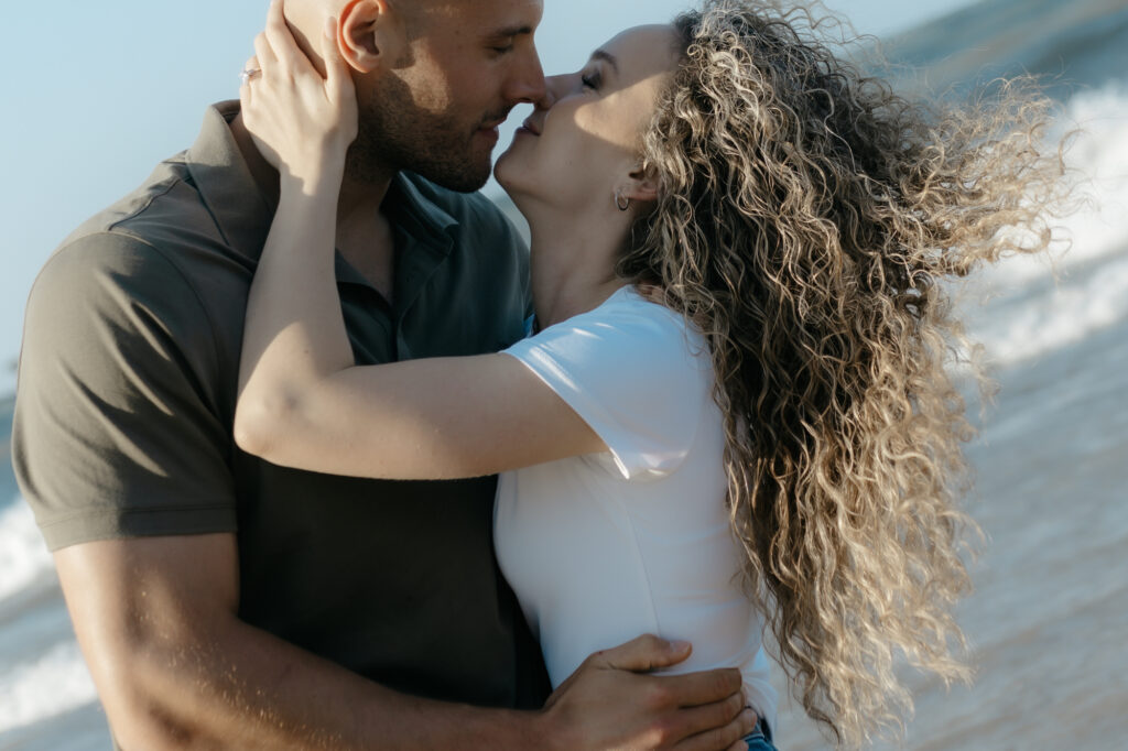 A close up of a couple kissing with wind blown curly hair during a session featuring fun engagement photo ideas.