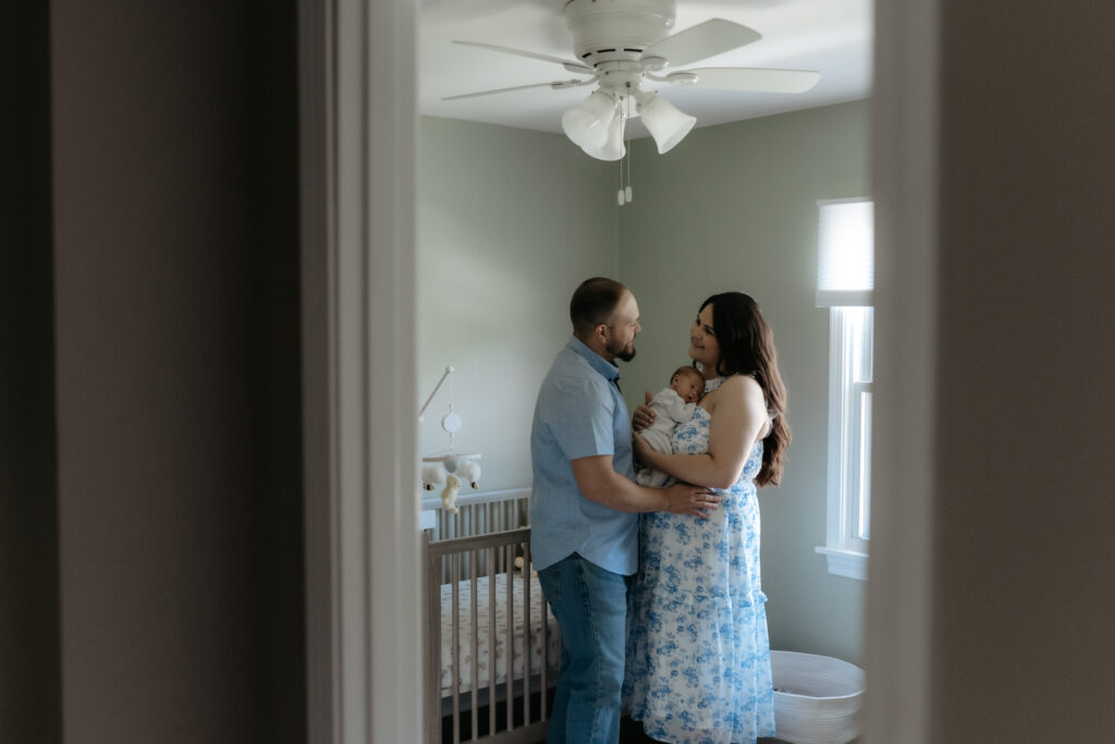 A mother and father hold their baby in a soft green nursery for at home newborn photography.