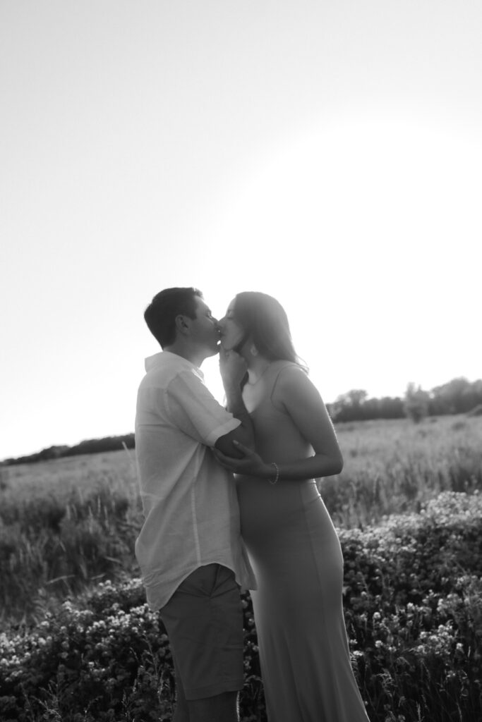 A romantic black and white photograph of a couple sharing an intimate kiss in a field of tall grass at sunset.