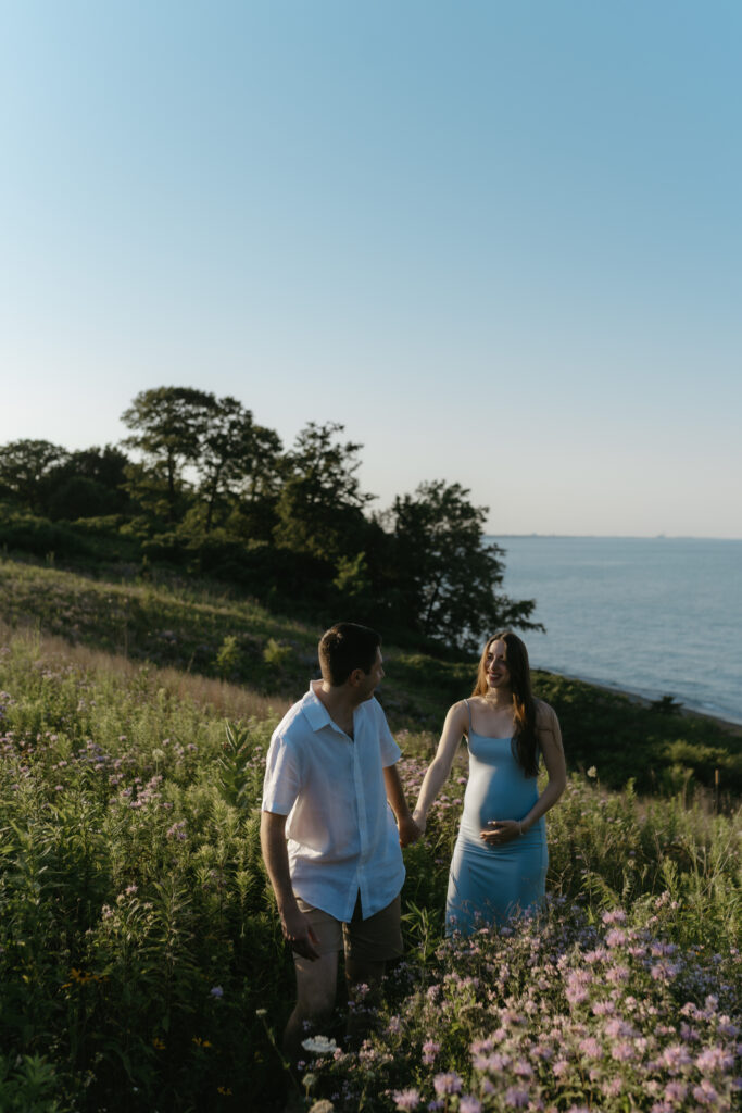 An expectant couple holds hands and walks through wildflowers to feel comfortable in front of the camera.