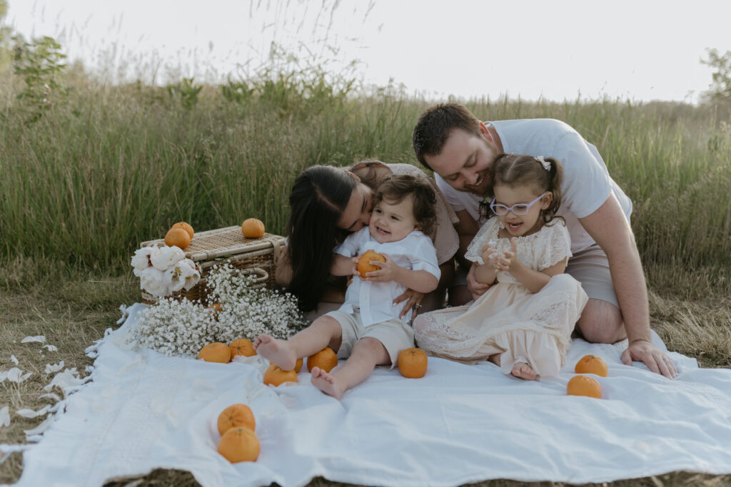 A mother and father smiling at their two young children during an outdoor maternity and family photo session on a picnic blanket.