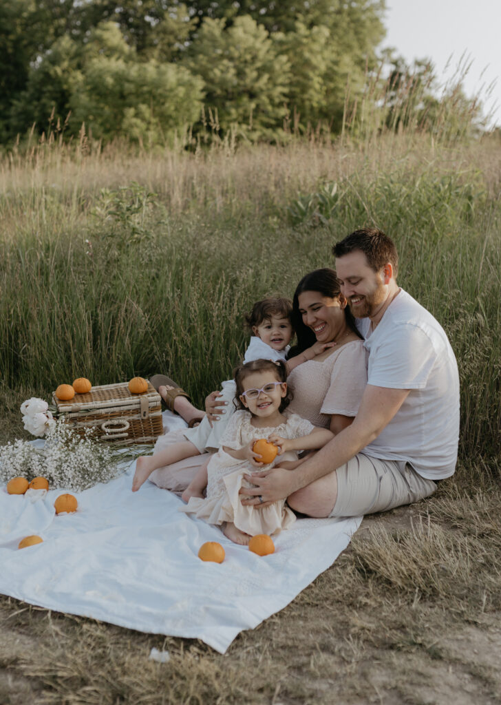 High-end family photo session in an outdoor grassy field with parents and two toddlers sitting on a white blanket during a styled picnic with oranges.