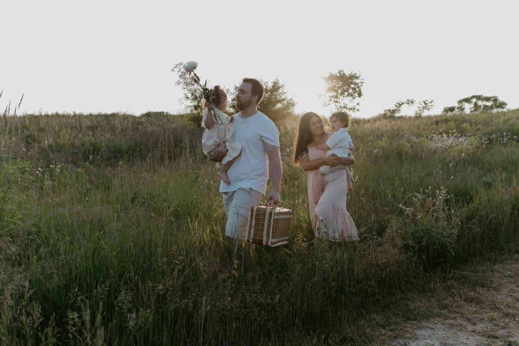 Parents walking through a tall grass field at sunset holding their toddlers and carrying a vintage wicker picnic basket.