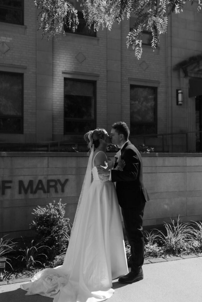 Black and white wedding portrait of a bride and groom kissing outside a stone building during their wedding ceremony.