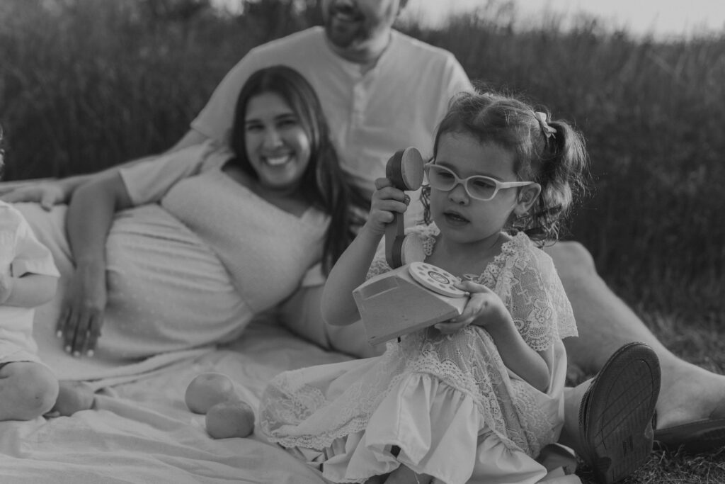 Black and white close up photo of a young girl wearing glasses playing with a vintage wooden rotary telephone toy during a family session.