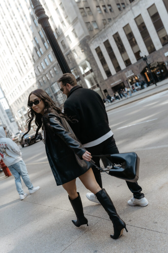 Candid movement shot of a stylish woman looking back over her shoulder in sunglasses and a black leather blazer crossing a city street.