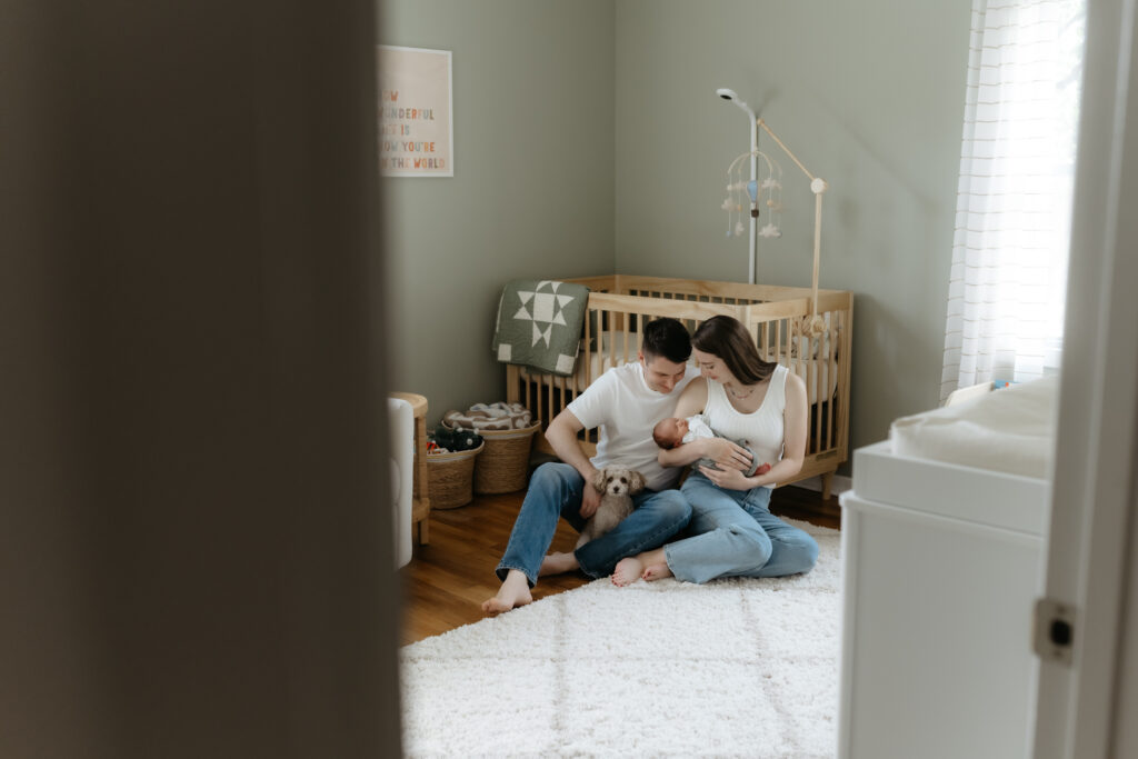 Parents sitting on a nursery rug cradling their newborn next to a wooden crib with their small dog.