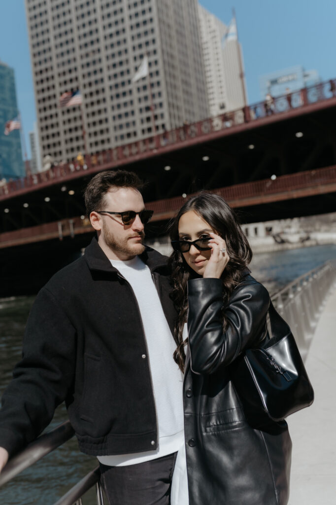 Fashion forward couple posing on a city riverwalk wearing black sunglasses and structured modern outfits.