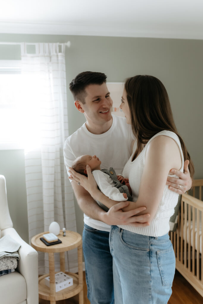Happy parents smiling at each other while holding their sleeping newborn baby in front of a nursery window.