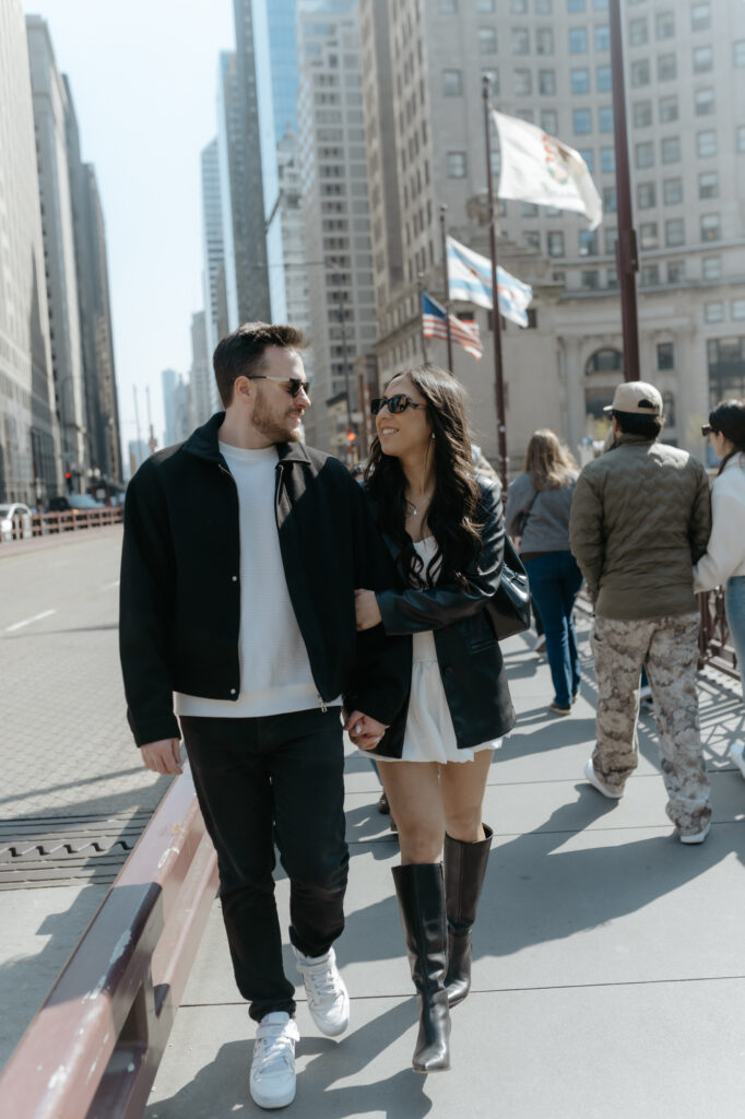 Chic engaged couple smiling and walking along a busy city bridge sidewalk during their editorial engagement session.