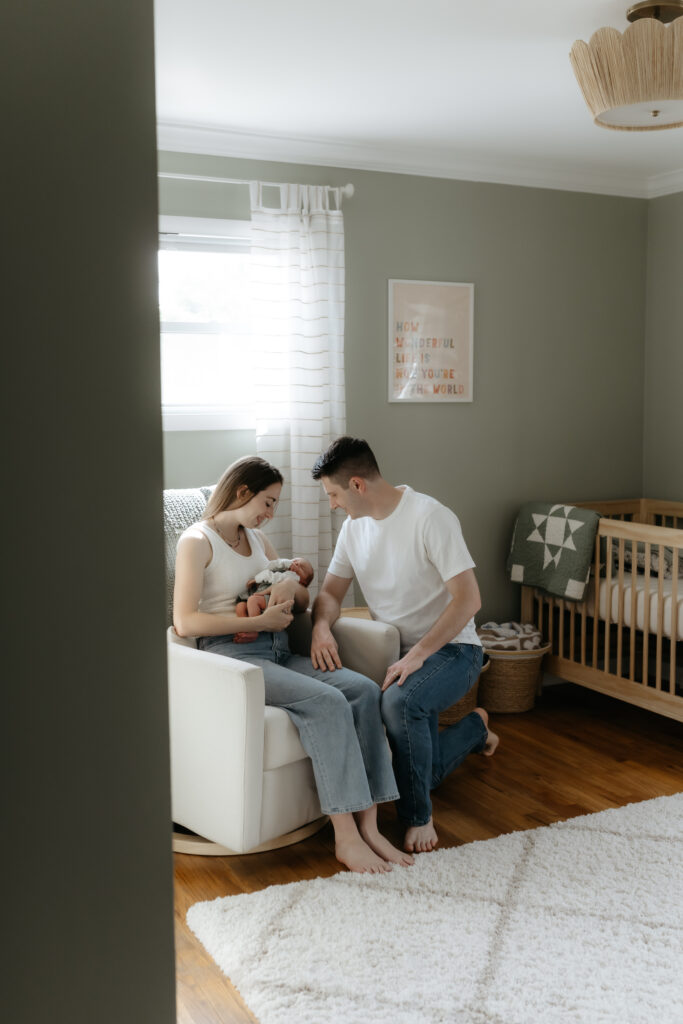 A mother holding her newborn baby in a white nursery rocking chair as the father kneels beside her.