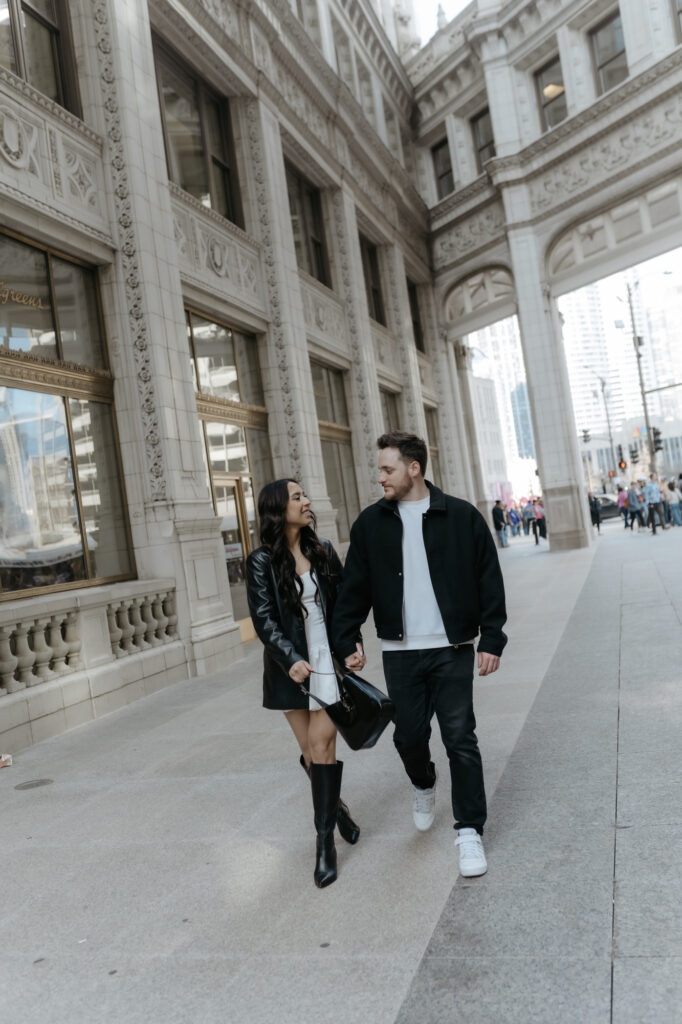 Engaged couple walking hand in hand past a historic white architectural building in downtown Chicago during a modern urban engagement session.