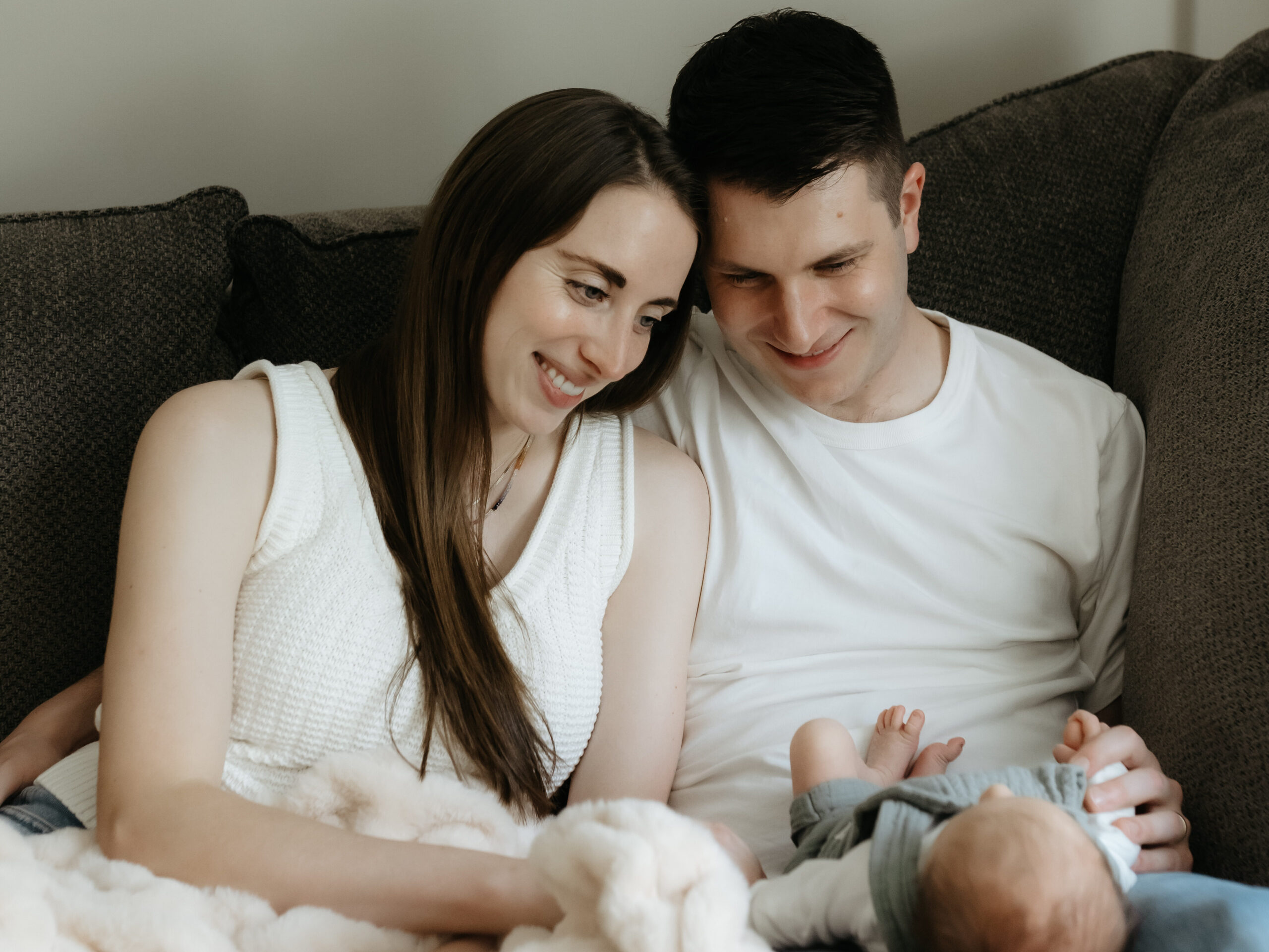 Parents lying on a couch smiling down at their sleeping newborn baby during an in-home session.