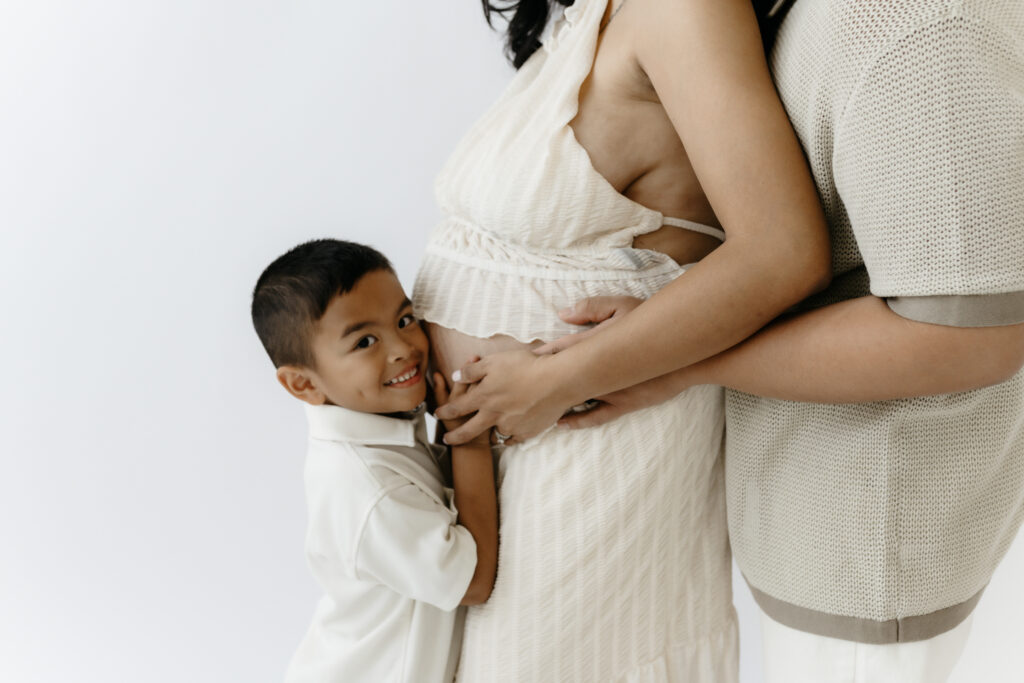 Close up detail shot of a young boy hugging his mother's pregnant belly while his father holds her waist in a white studio.
