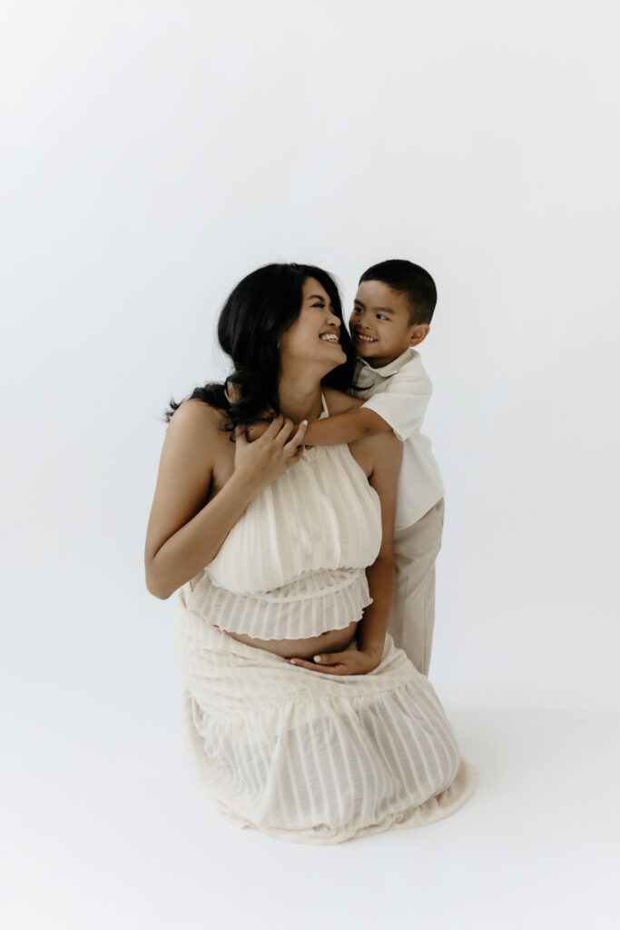 A smiling pregnant mother kneeling on a white studio floor while her young son hugs her from behind during a studio maternity session.