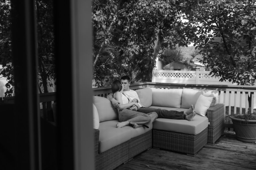 Black and white photo of parents relaxing on an outdoor deck couch cuddling with their newborn baby.