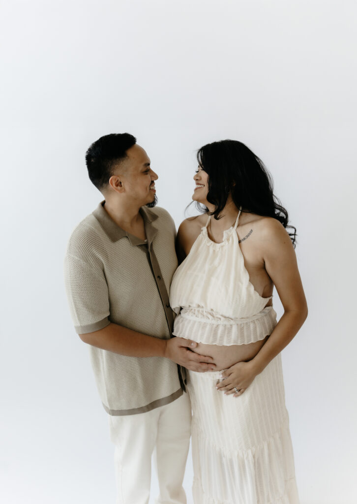 An elegant couple looking at each other and smiling during an editorial studio maternity photo shoot against a plain white background.