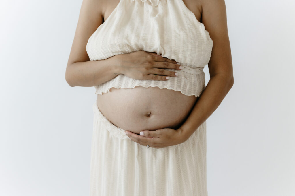 Midsection portrait of a pregnant woman in a cream pleated crop top and skirt holding her bare belly in front of a white backdrop.