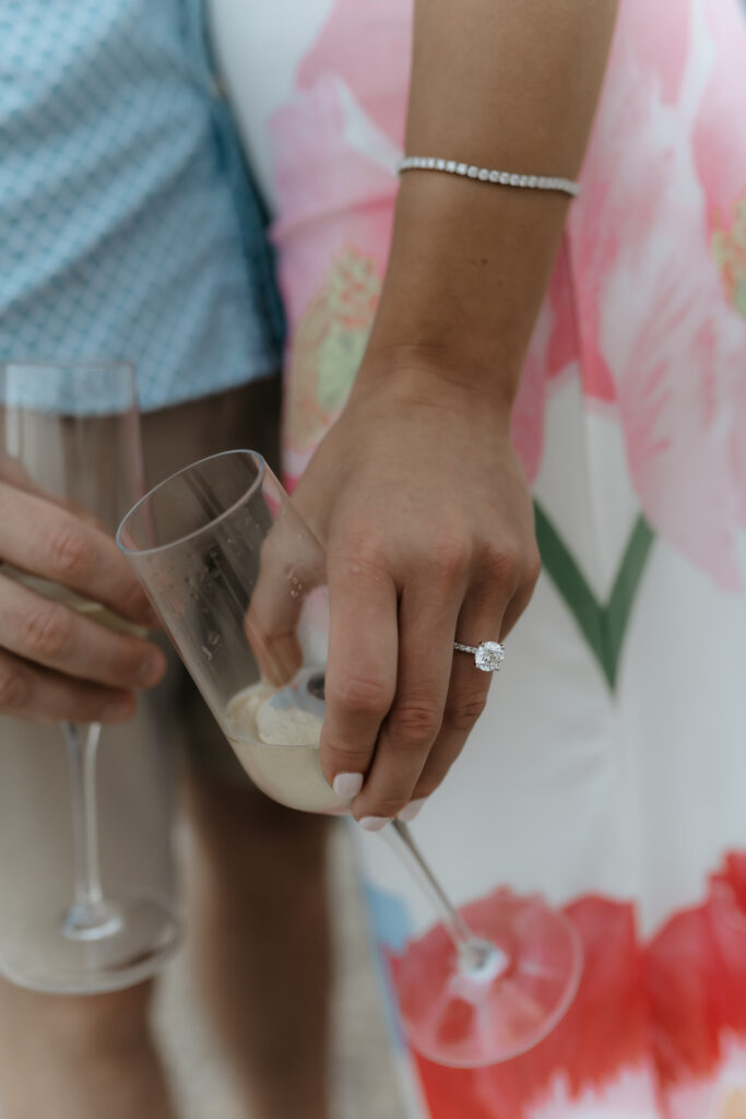 Close-up detail of a bride wearing a diamond engagement ring and tennis bracelet while holding a glass of champagne with her fiancé.