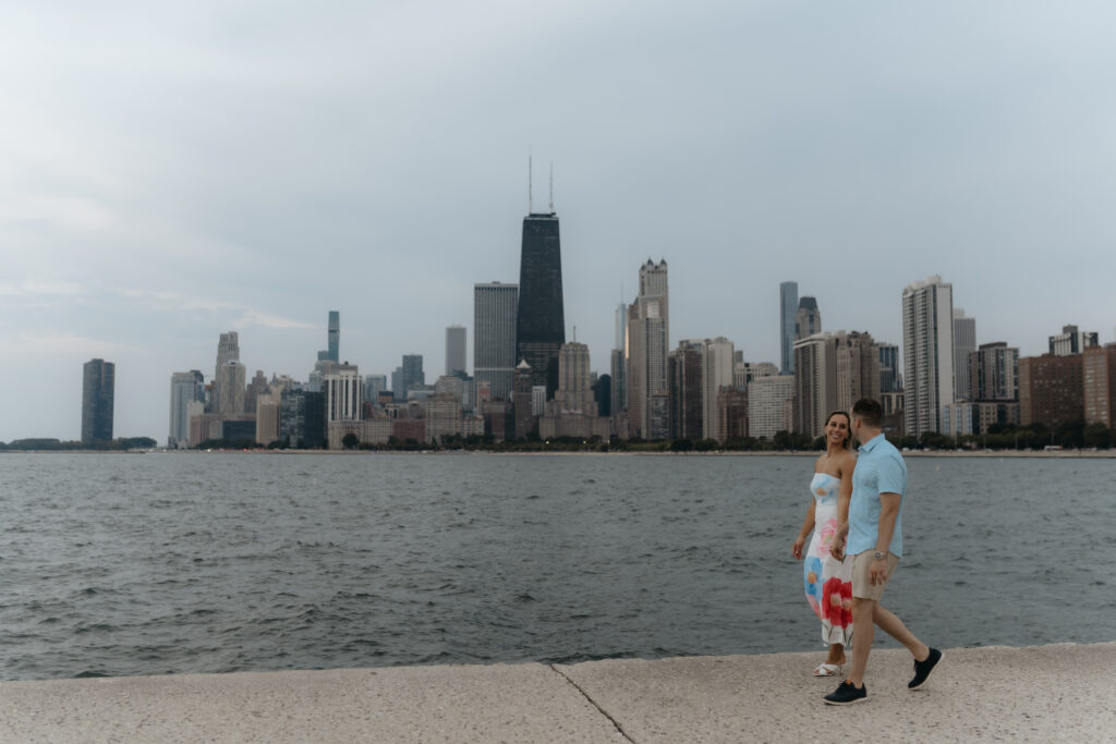 An engaged couple holding hands and walking along a concrete pier with the towering Chicago skyline and Lake Michigan in the background.
