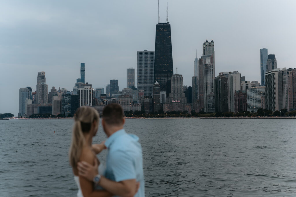 Out of focus shot of a couple embracing with a clear, sharp view of the John Hancock building and Chicago skyline across the water.