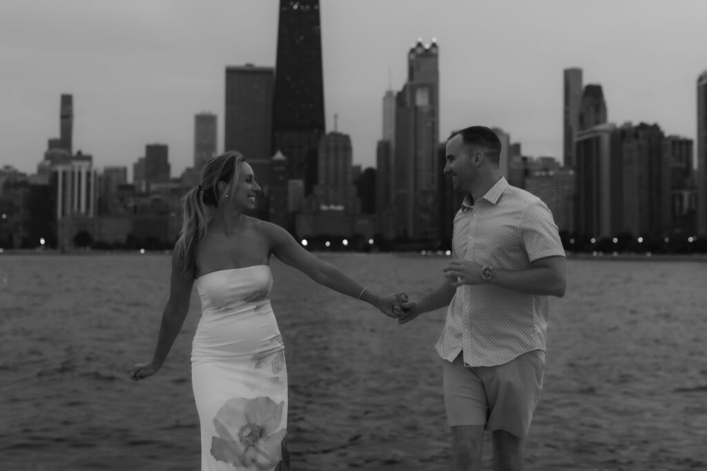 Black and white portrait of a happy couple holding hands and running along the lakefront with the city skyline behind them.