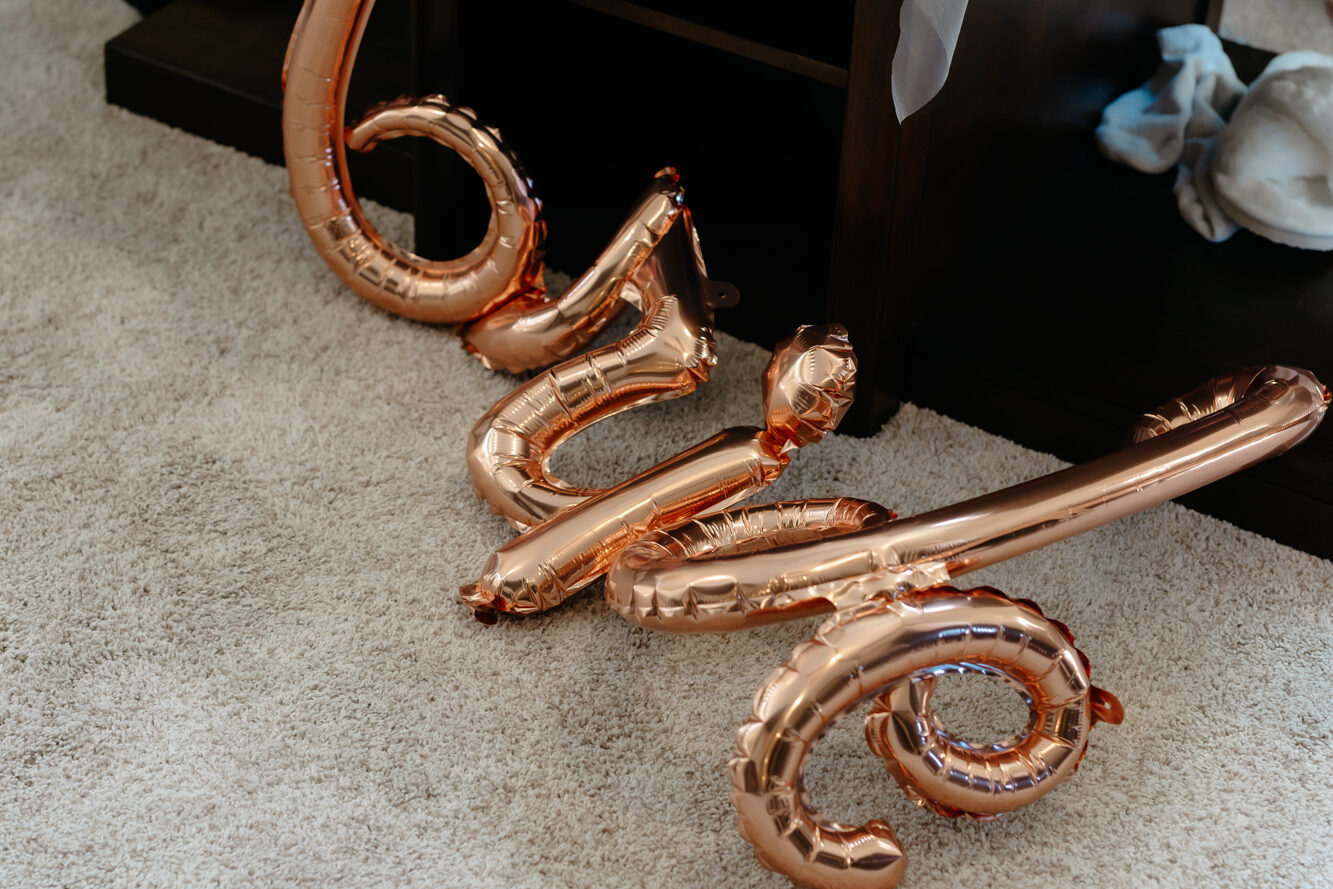 Rose gold metallic balloons spelling out love on a beige carpet next to a coffee table with bridal morning prep items.