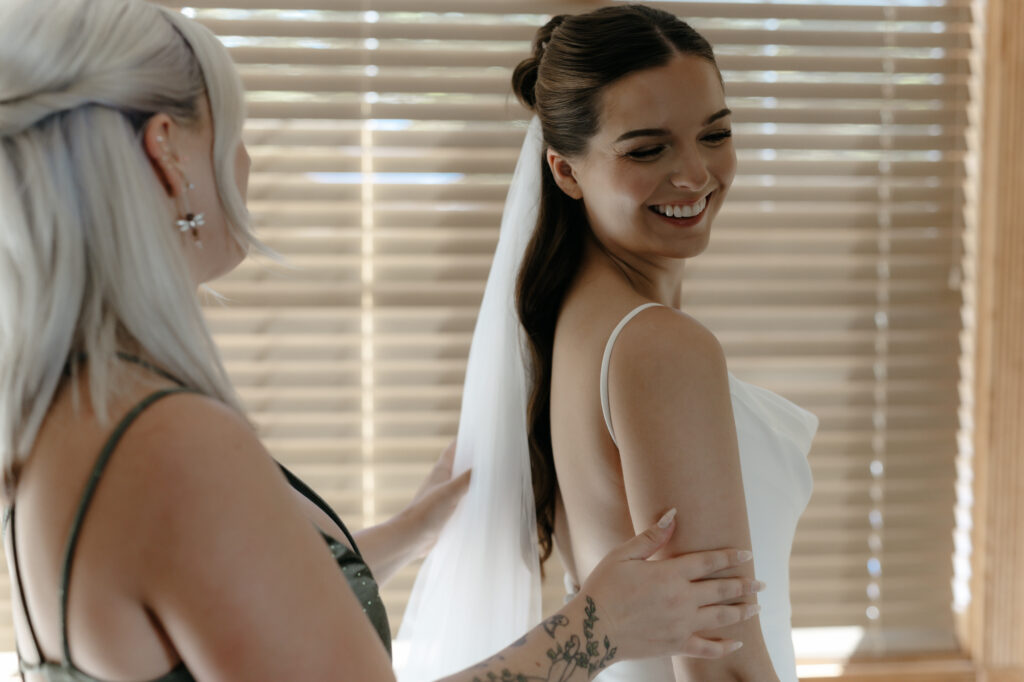 Candid shot of a bridesmaid adjusting a bride's wedding veil as the bride laughs happily during morning preparation.