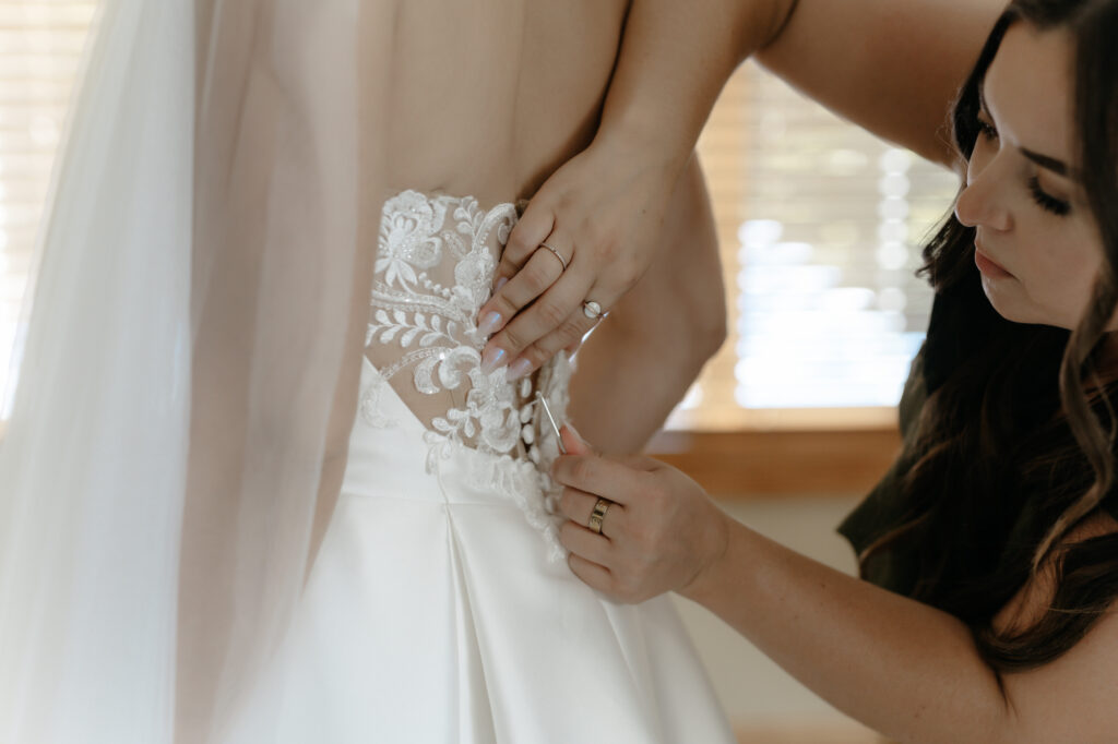 Close-up detail shot of a bridesmaid using a button hook to fasten the delicate lace back of a bride's wedding dress.