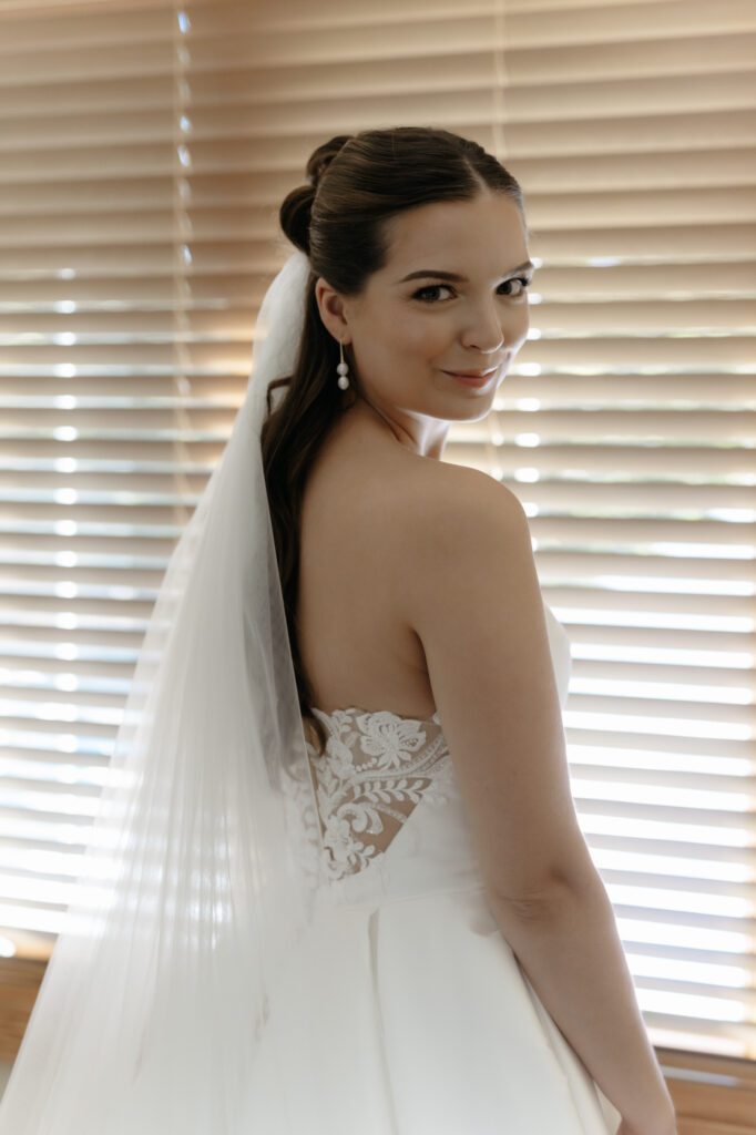 Editorial over-the-shoulder portrait of a bride smiling in her strapless wedding gown with a classic veil and pearl earrings.