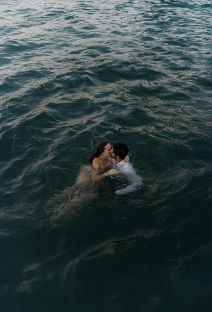 Editorial portrait of an engaged couple swimming in the ocean while wearing their formal attire during a sunset session.
