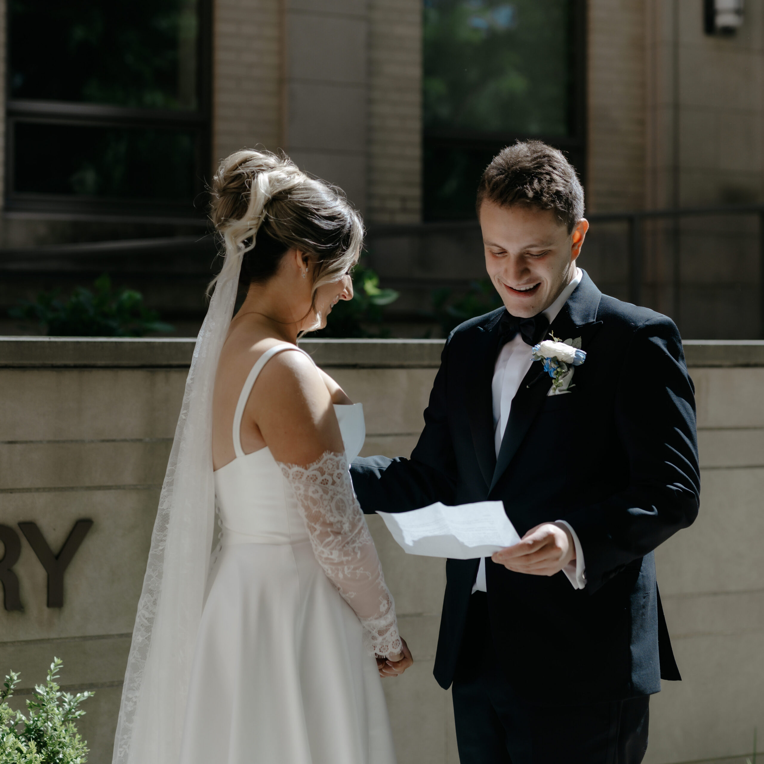 A groom laughing joyfully while reading his wedding vows to his bride during an outdoor ceremony.