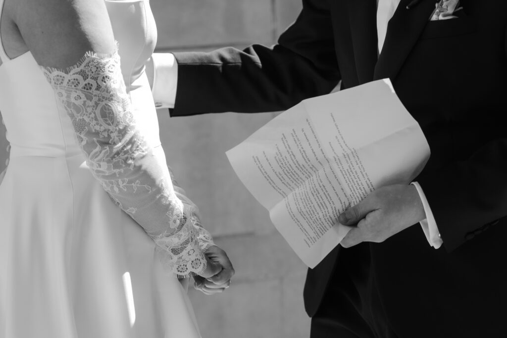 Close-up black and white shot of a groom holding his printed wedding vows next to his bride wearing lace sleeves.