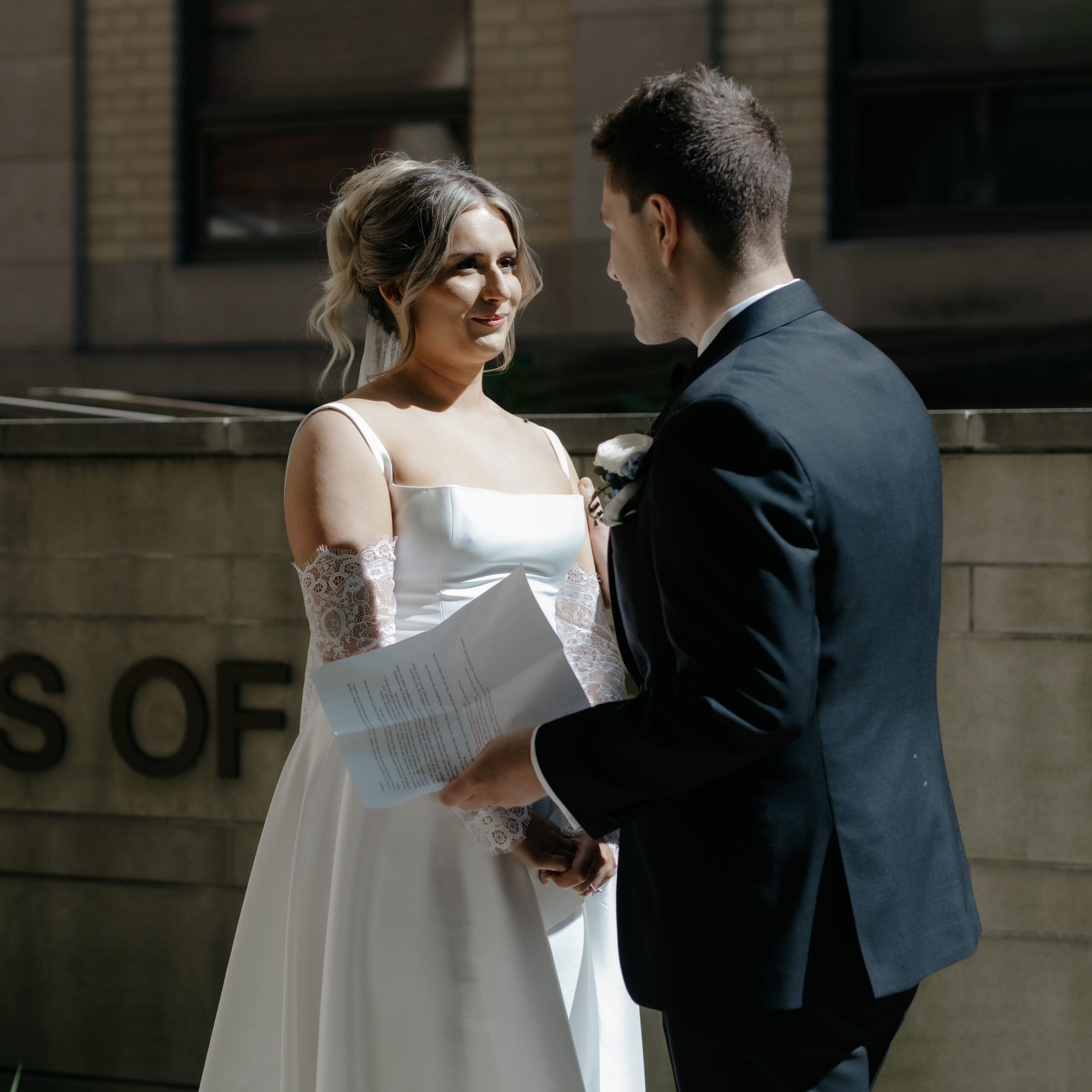 A bride wearing a satin gown and lace sleeves looking lovingly at her groom as he holds his wedding vows.