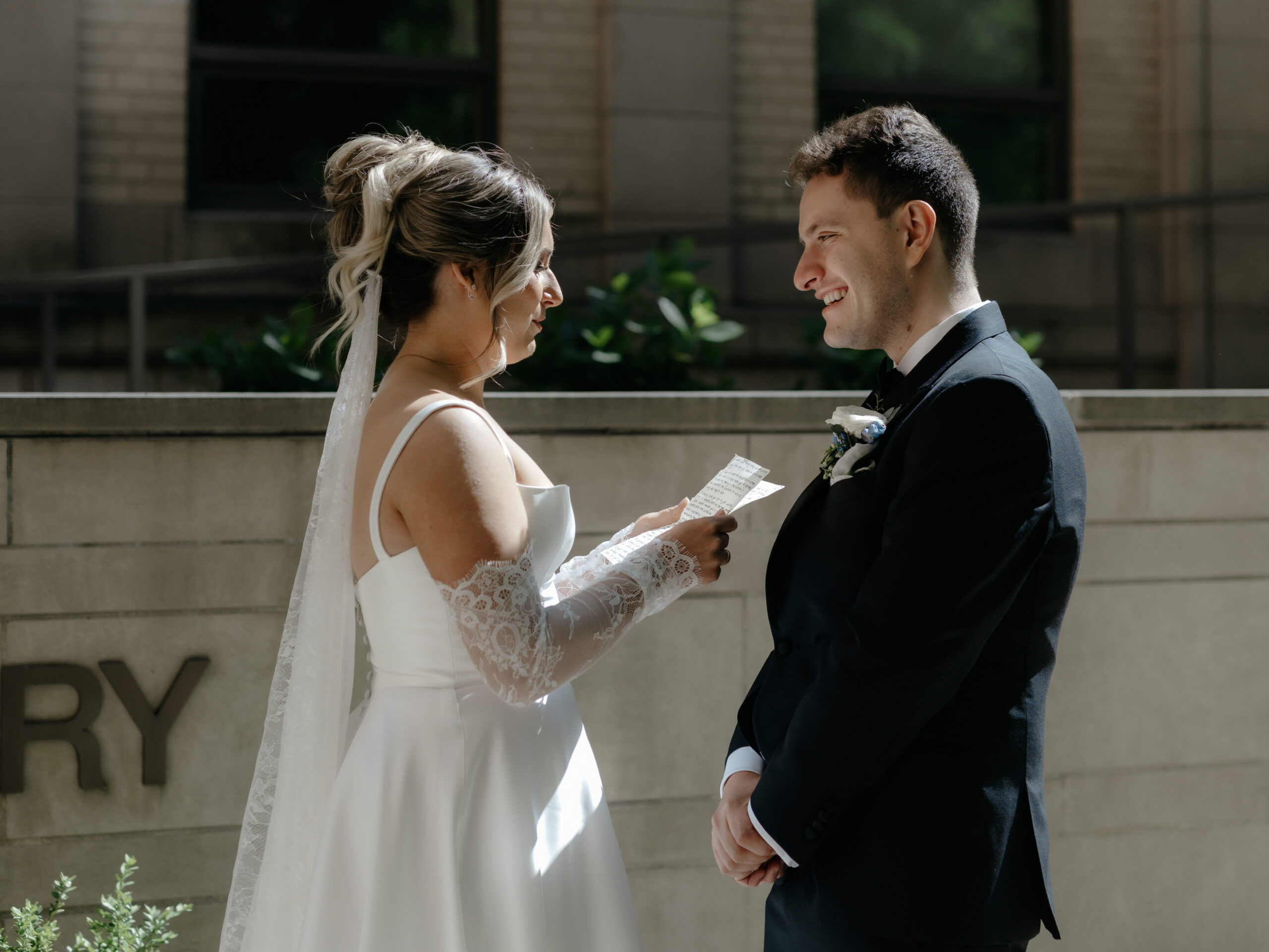 A groom smiling happily at his bride as she reads her wedding vows to him outside a brick building.