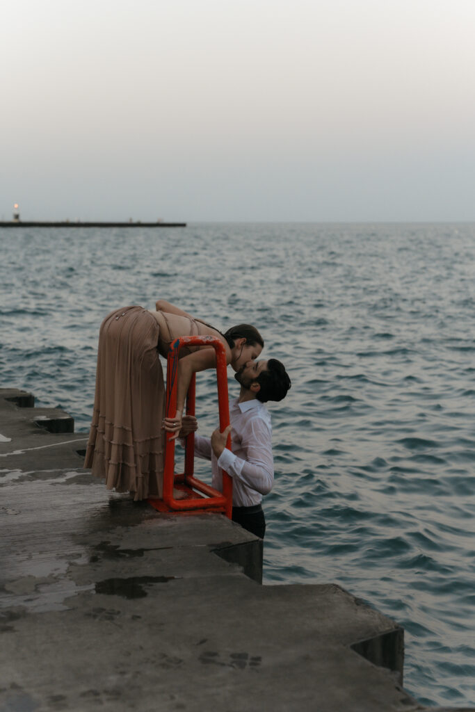 Cinematic shot of a woman leaning over an orange ladder on a pier to kiss her fiancé who is standing in the water.